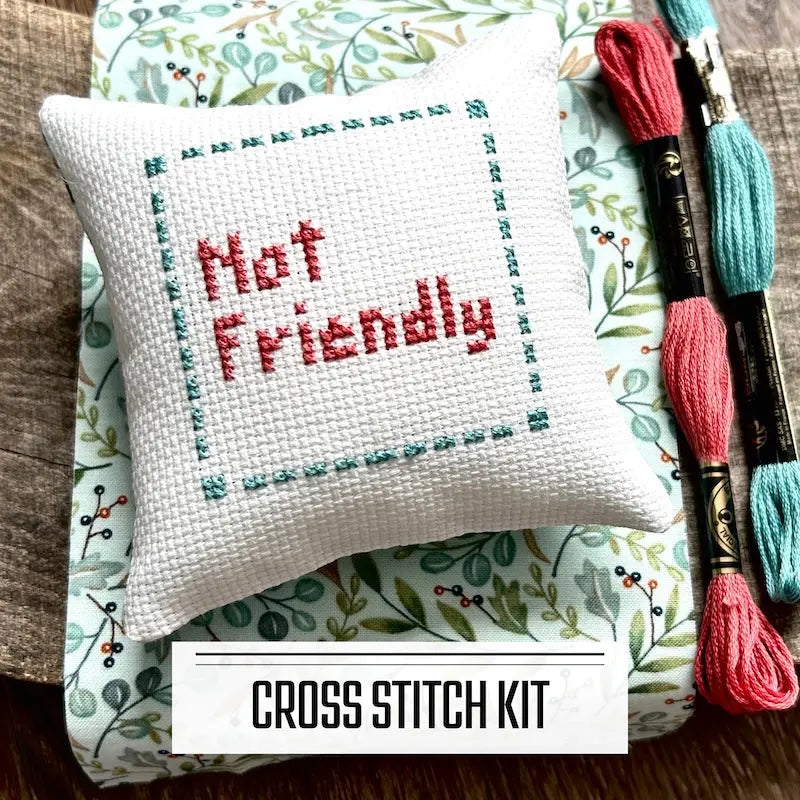 A DIY cross stitch kit for a mini pillow that can be used as a pin cushion. The finished pillow is sitting on a wooden board.