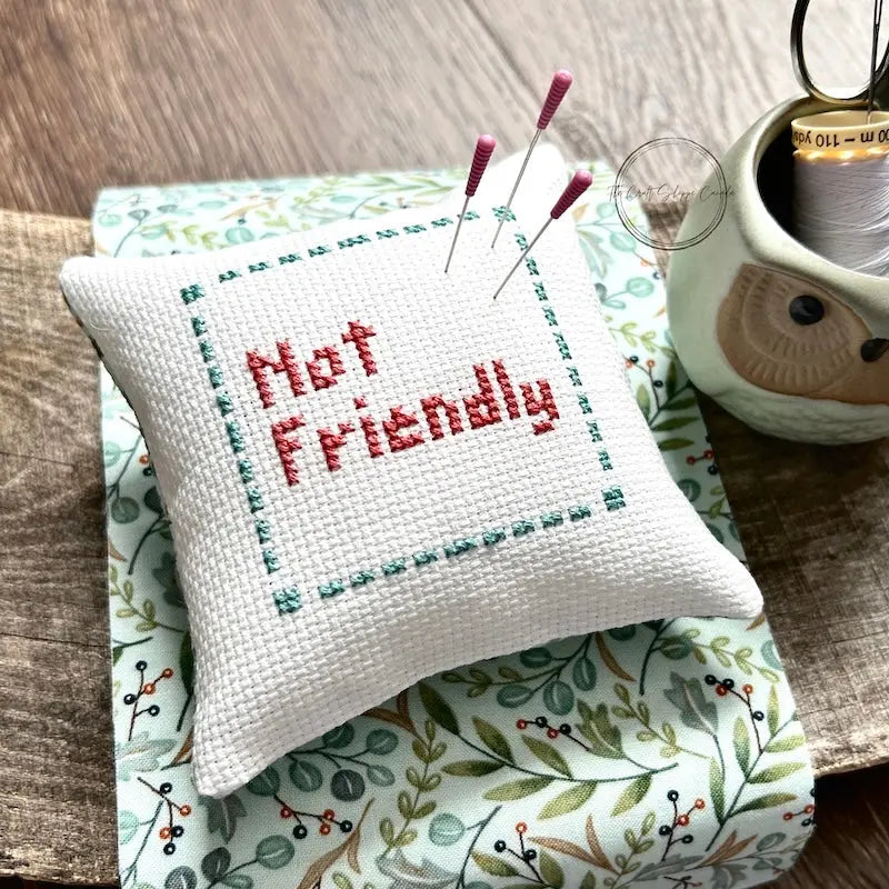 The finished pillow, made from this cross stitch pattern, is sitting on a wooden board with pins sticking out.