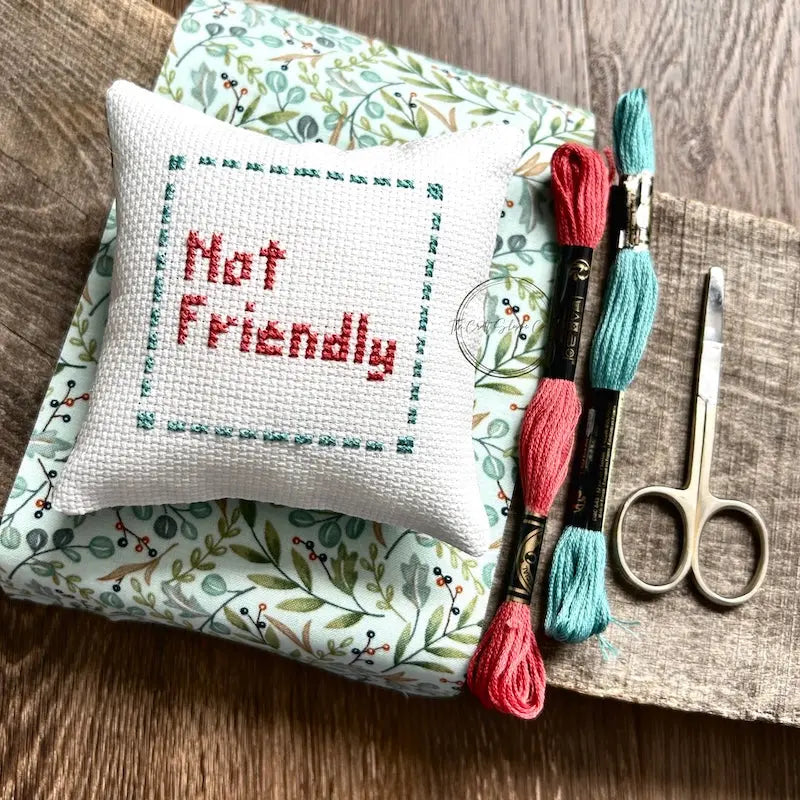 The finished pillow, made from this cross stitch kit, is sitting on a board with all of the supplies needed.