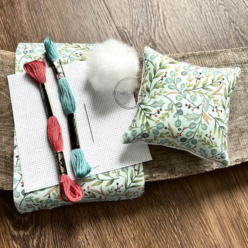 The supplies needed to make this cross stitch mini pillow are sitting on a wooden board.