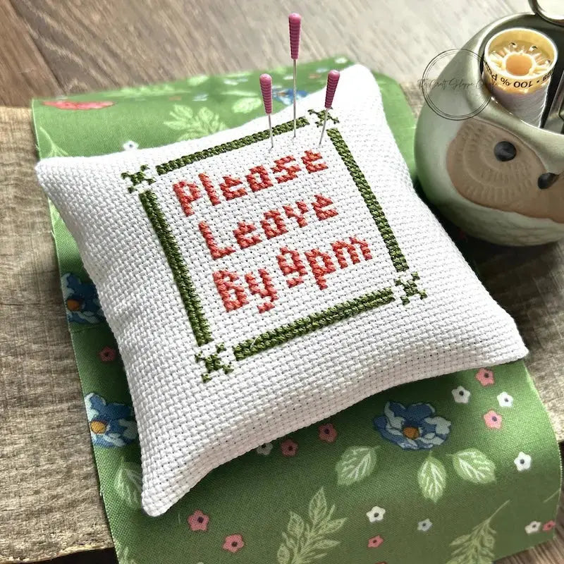 The finished pillow, made from this cross stitch pattern, is sitting on a wooden board with pins sticking out.