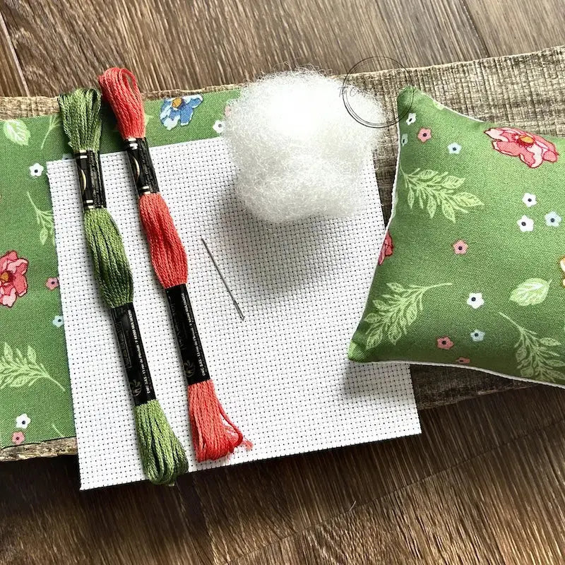 The supplies needed to make this cross stitch mini pillow are sitting on a wooden board.