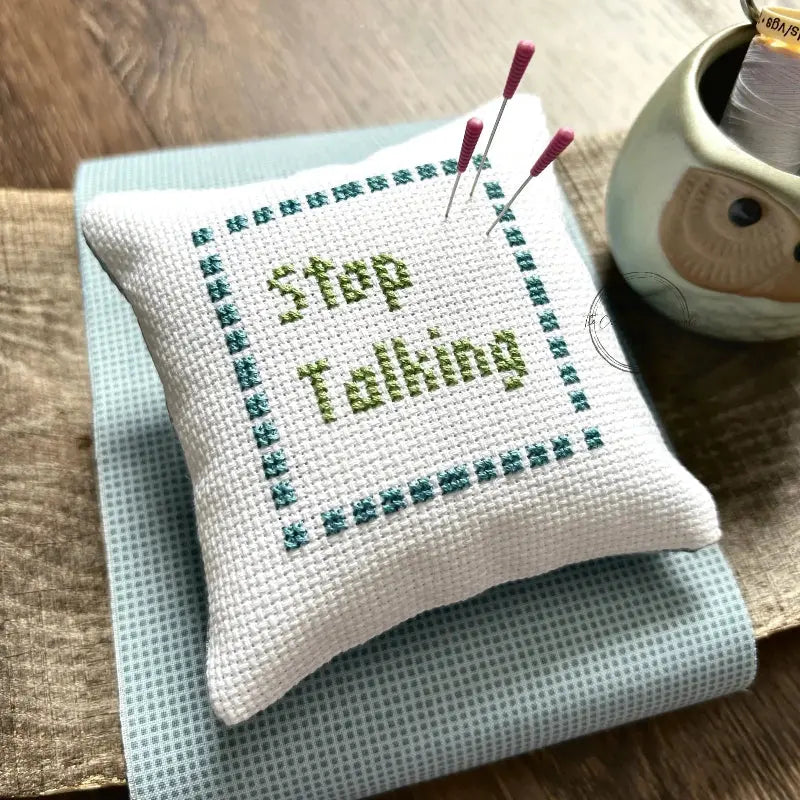 The finished pillow, made from this cross stitch pattern, is sitting on a wooden board with pins sticking out.