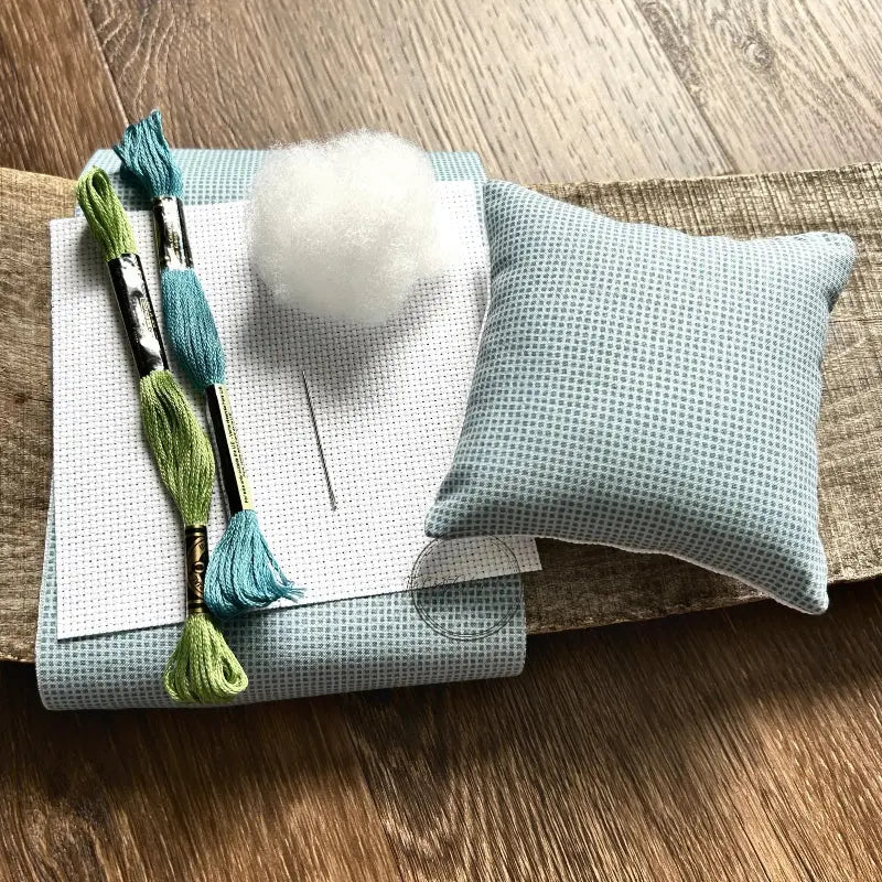 The supplies needed to make this cross stitch mini pillow are sitting on a wooden board.