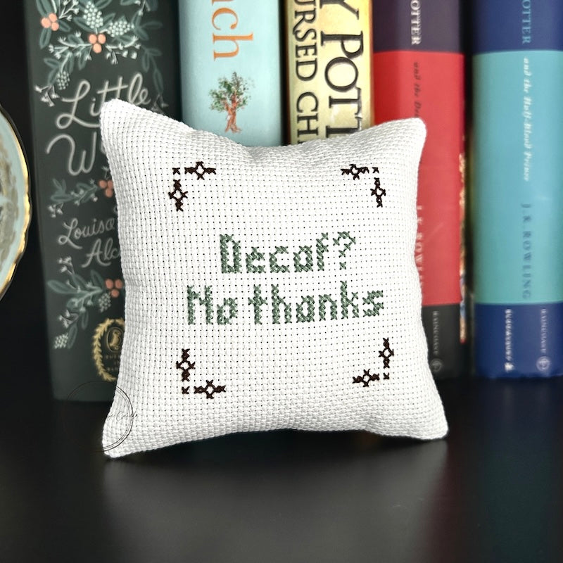 The finished pillow, made from this cross stitch kit, is sitting on a book shelf.