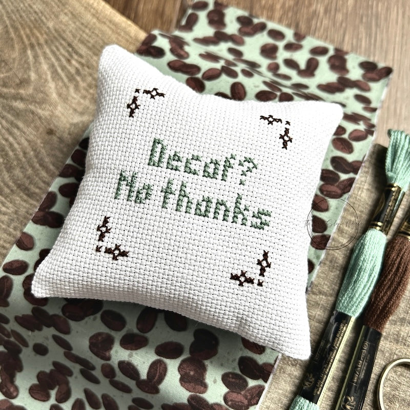 The finished pillow, made from this cross stitch kit, is sitting on a board with all of the supplies.