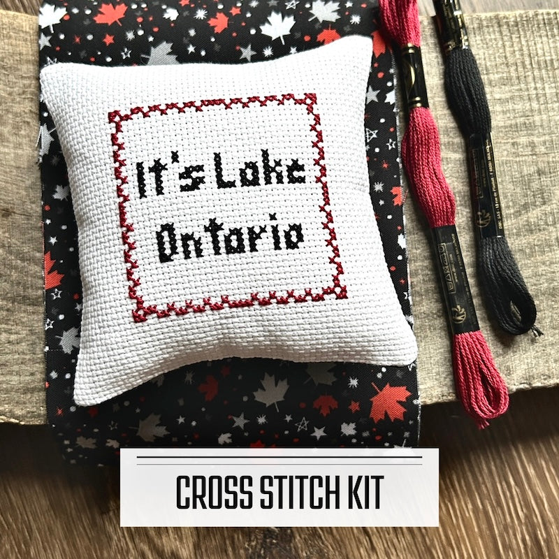 A DIY cross stitch kit for a mini pillow that can be used as a pin cushion. The finished pillow is sitting on a wooden board.