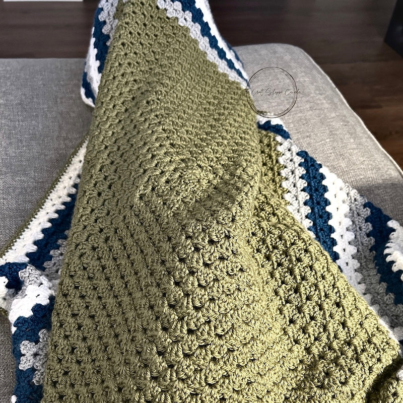 The finished green, teal and grey blanket laying over someone's lap on a couch - The Craft Shoppe Canada