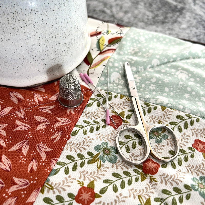 The finished hot pad sitting on a wooden board with a ceramic mug on top. There are scissors, pins and a thimble laying on top - The Craft Shoppe Canada