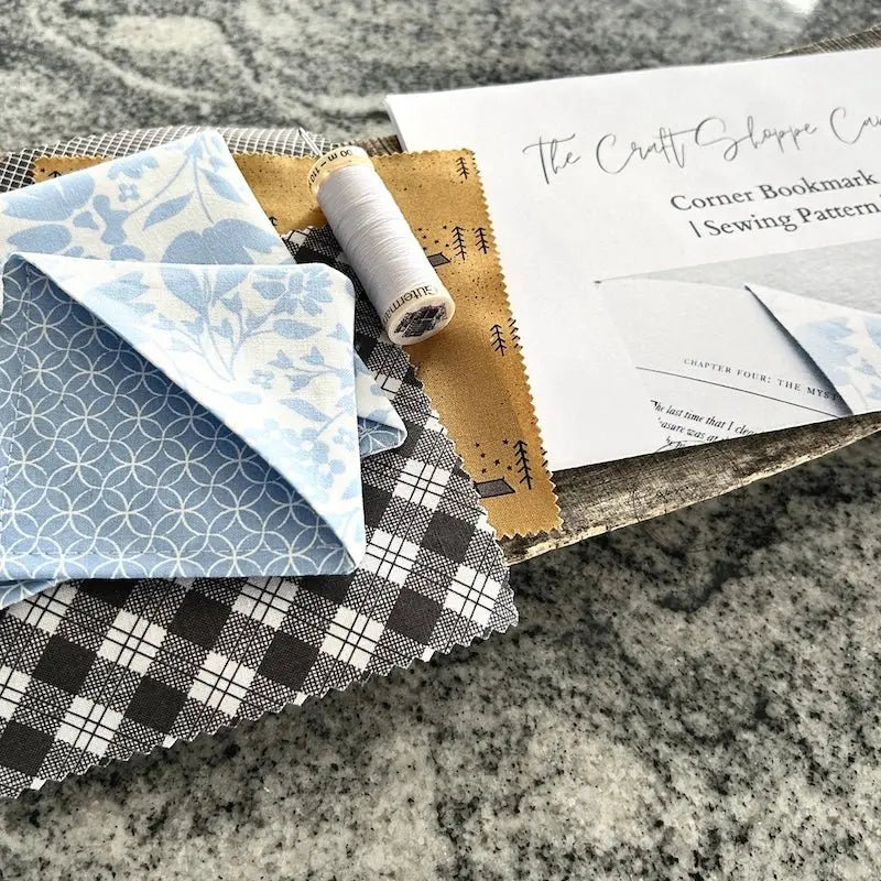 The corner bookmarks, from this sewing kit, are sitting on a wooden board with the sewing pattern, a spool of white thread and a hand sewing needle.