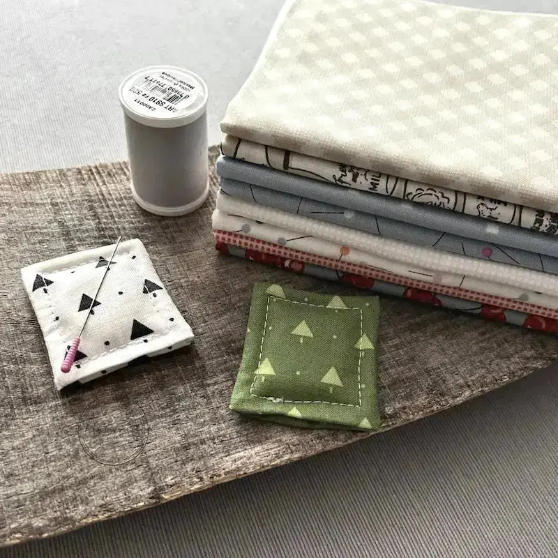 The 2 needle minders, made from this sewing kit, are sitting on a wooden board beside a stack of fabric, and a spool of thread.