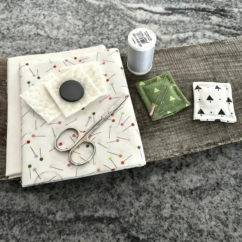 The 2 needle minders, made from this sewing pattern, are sitting on a wooden board beside a stack of fabric, a magnet, cotton batting and a spool of thread.