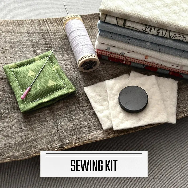 A DIY sewing kit for a needle minder/fridge magnet. There are 2 needle minders lying on a wooden board with fabric, a spool of thread and a magnet.