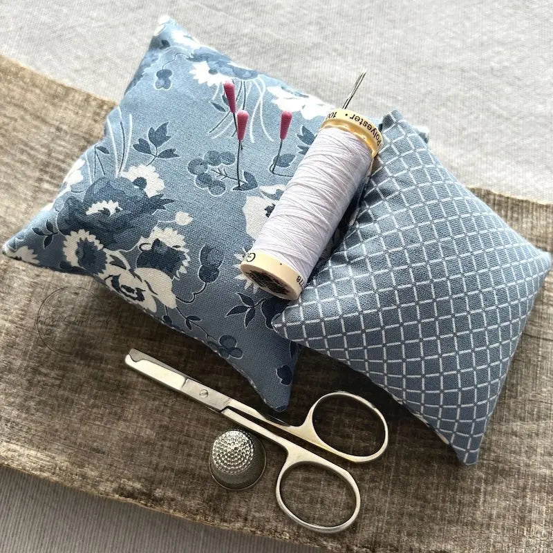 The pin cushions, from this sewing kit are sitting on a wooden board with a pair of scissors, a thimble, a spool of white thread and a sewing needle.