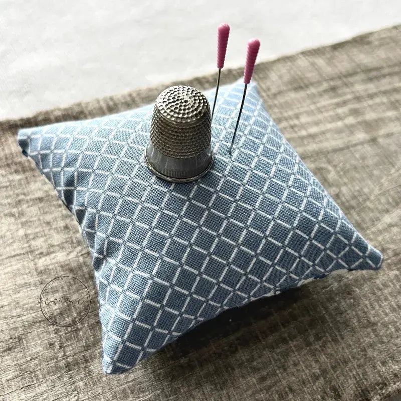 A pin cushion, from this sewing kit, sitting on a wooden board with a thimble and sewing pins on top.