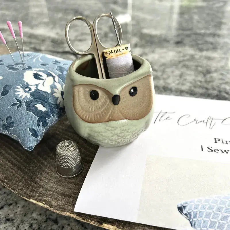 A pin cushion, from this sewing kit, is sitting on a wooden board with sewing pins, scissors, a copy of the sewing pattern and a spool of white thread.