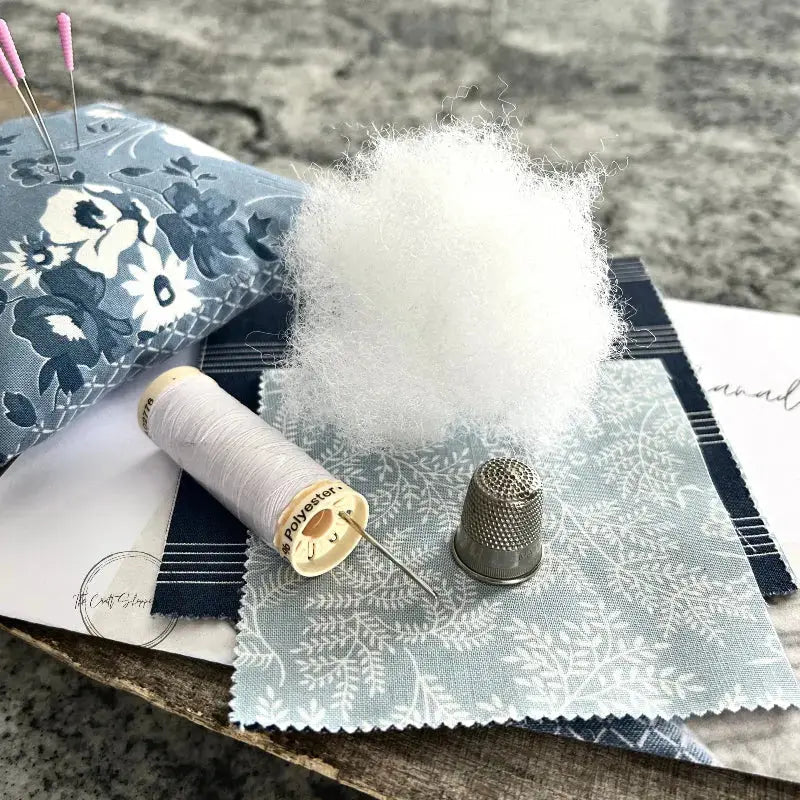 A pin cushion, from this sewing kit, is sitting on a wooden board with fabric, poly fil stuffing, scissors, a copy of the sewing pattern and a spool of white thread.