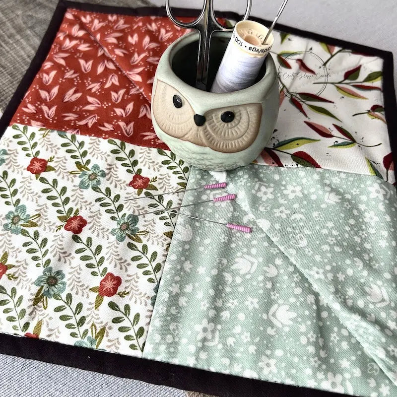 The pot holder trivet, made from this sewing pattern, is sitting on a wooden board with a pair of scissors, a spool of thread, and sewing pins.