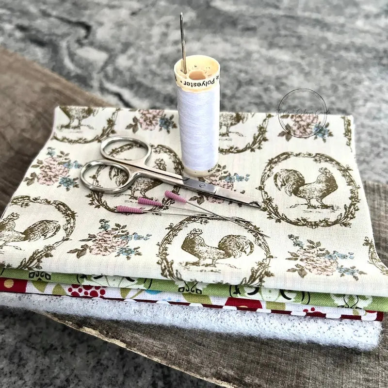 Fabric used to make the hot pad, from this sewing pattern, is sitting on a wooden board with a pair of scissors, a spool of thread, and sewing pins.