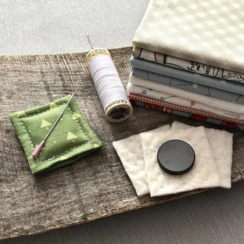 A needle minder, made from this sewing pattern, is sitting on a wooden board beside a stack of fabric, cotton batting, a magnet and a spool of thread.