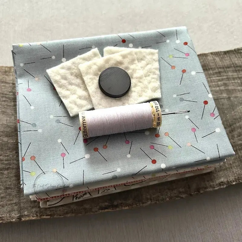 A stack of fabric, cotton batting and a magnet, that would be used in this sewing pattern, are sitting on a wooden board with a spool of thread and a hand sewing needle.