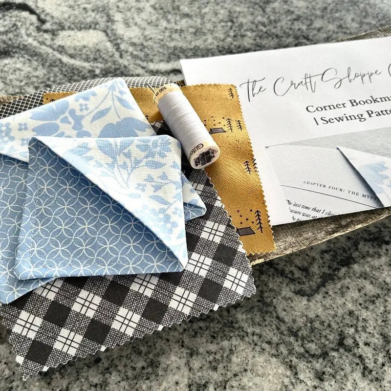The sewing pattern is laying on a wooden board with fabric, the finished bookmarks, a spool of white thread and a sewing needle beside it