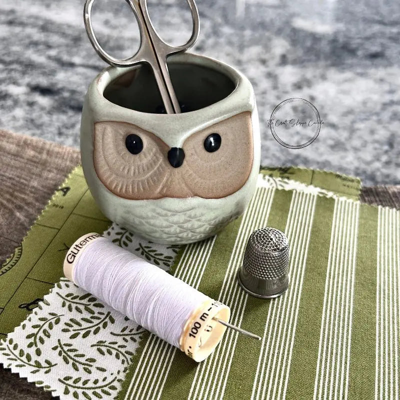 Fabric that would be used in this sewing pattern, is sitting on a wooden board with a spool of thread, a hand sewing needle, scissors and a thimble.
