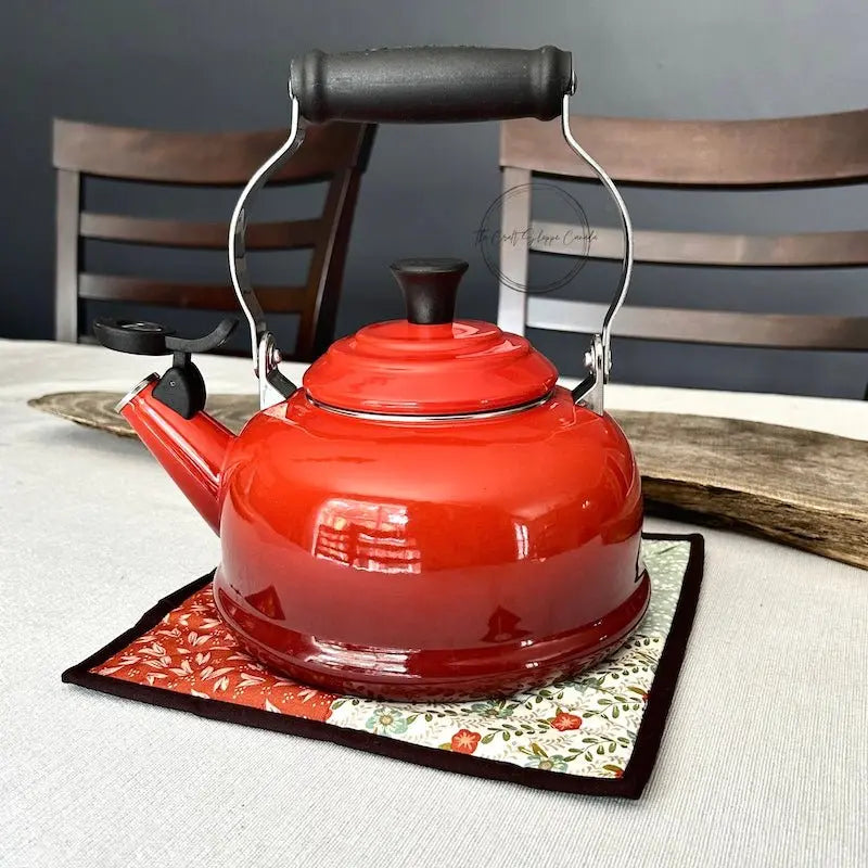 The finished hot pad, made from this sewing pattern, is sitting on top of a table with a red kettle on top.