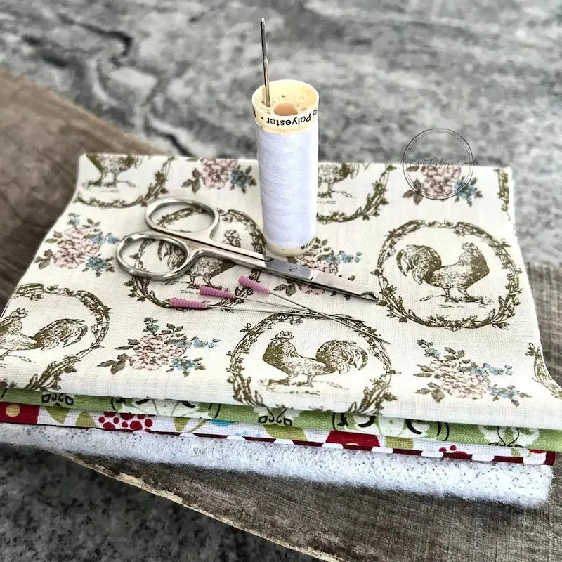 Fabric used to make the hot pad, from this sewing pattern, is sitting on a wooden board with a pair of scissors, a spool of thread, and sewing pins.