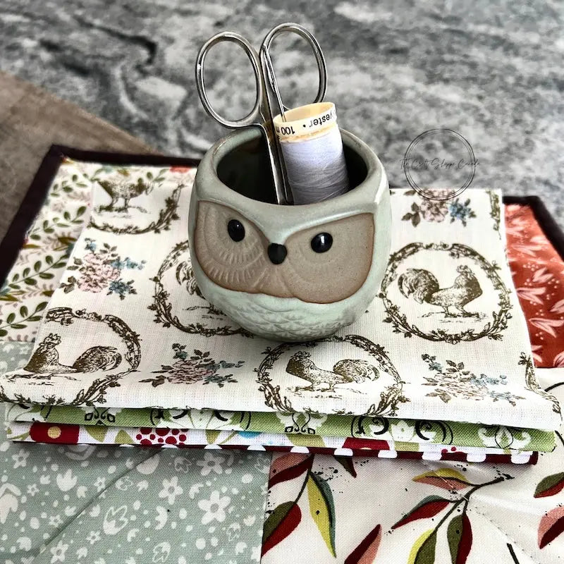 Fabric used to make the hot pad, from this sewing pattern, is sitting on a wooden board with a pair of scissors, a spool of thread, and sewing pins.