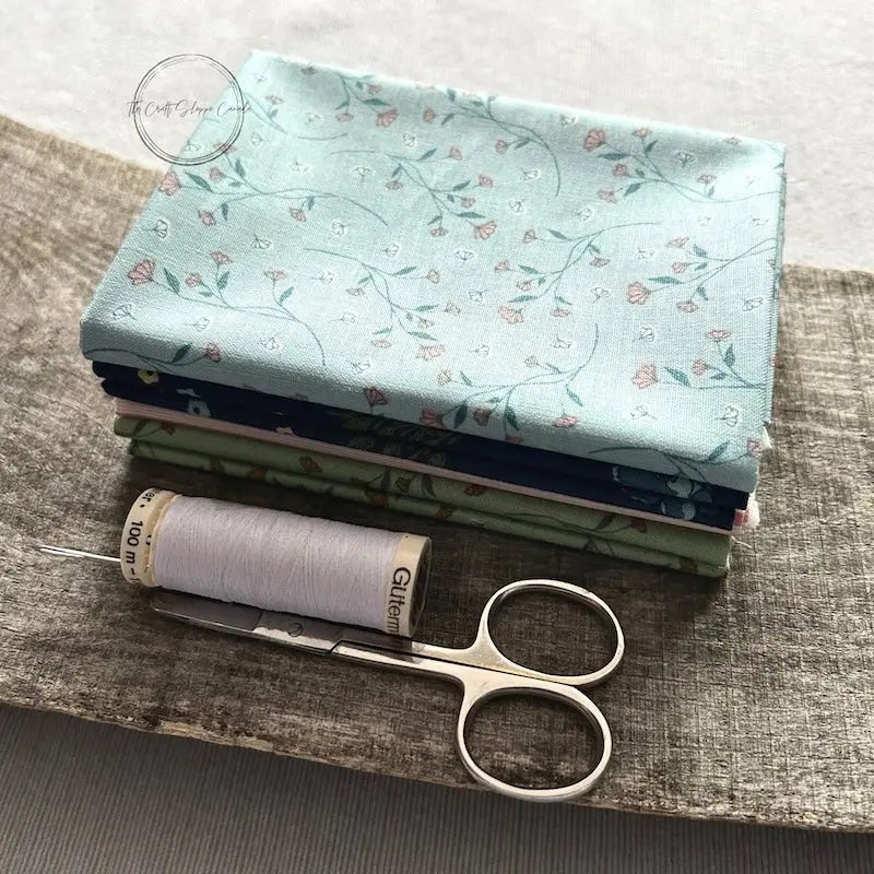 A pile of fabric that would be used for this sewing pattern, is lying on a wooden board with a spool of white thread and a hand sewing needle.