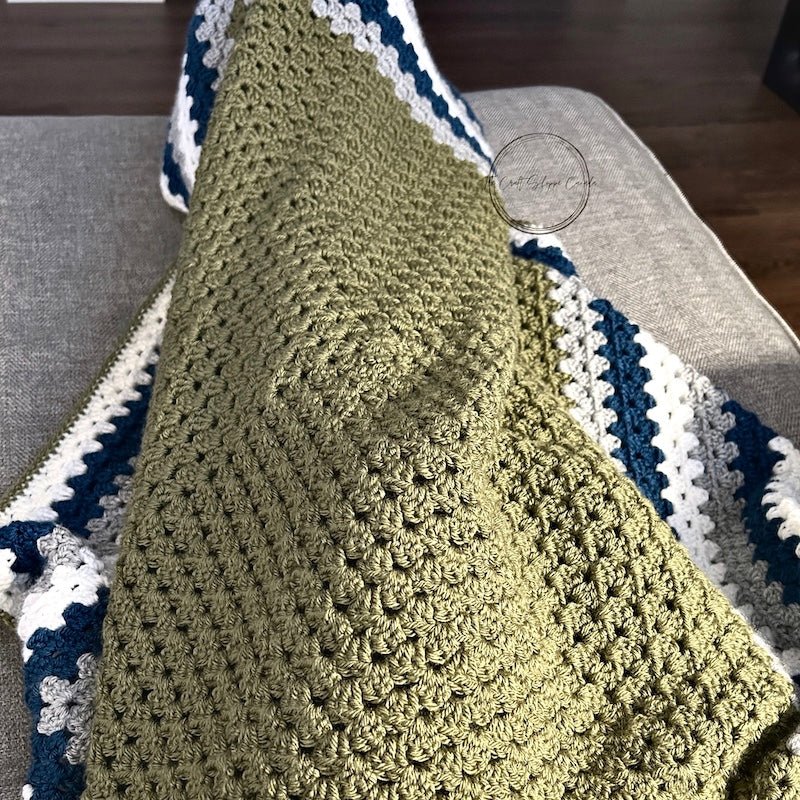 The finished green, teal and grey blanket laying over someone's lap on a couch - The Craft Shoppe Canada