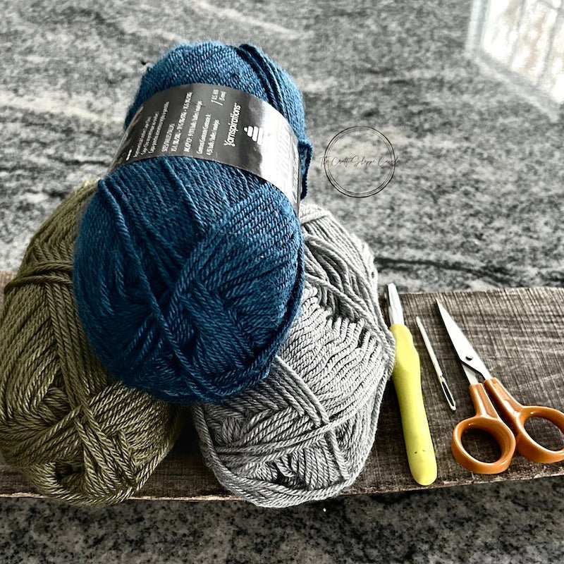3 balls of yarn on a wooden board with a crochet hook, a tapestry needle and a pair of scissors - The Craft Shoppe Canada