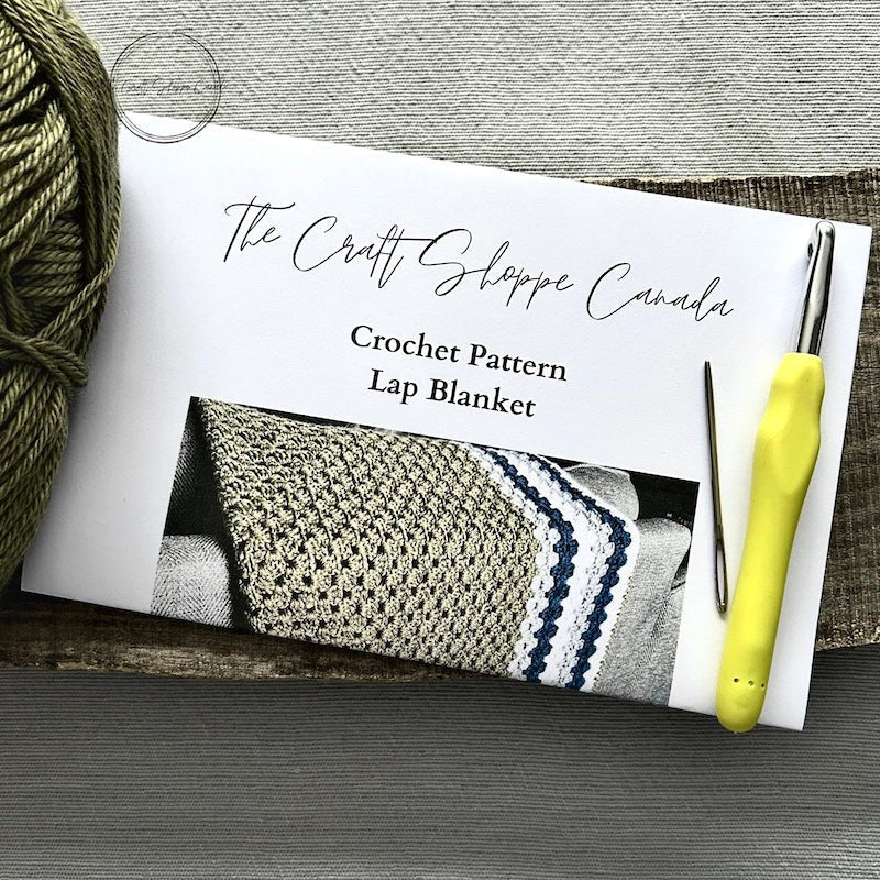 A copy of the lap blanket crochet pattern with a crochet hook, ball of yarn, and a tapestry needle - The Craft Shoppe Canada