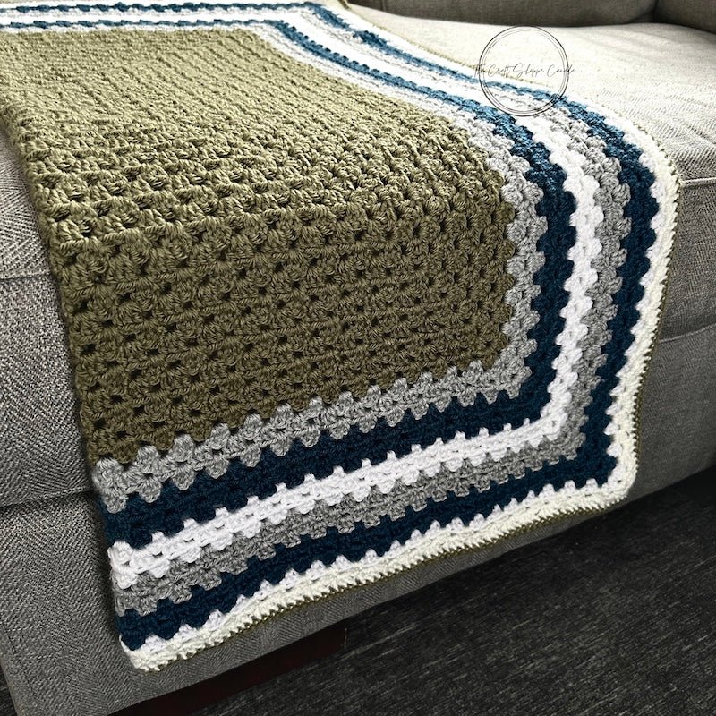 The finished green, teal and grey blanket hanging over a stool from the couch - The Craft Shoppe Canada