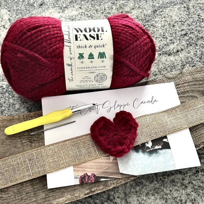 A finished heart, made from this crochet pattern, is lying on top of the pattern with a ball of red yarn, a crochet hook and a tapestry needle.