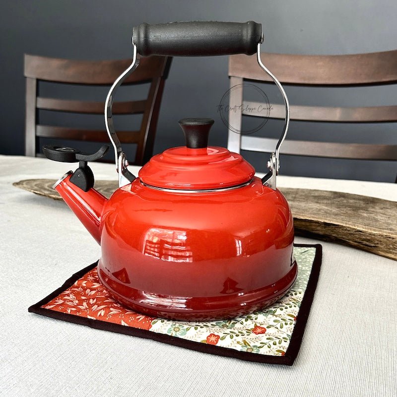 The finished hot pad is sitting on top of a table. A red kettle is on top of the hot pad - The Craft Shoppe Canada