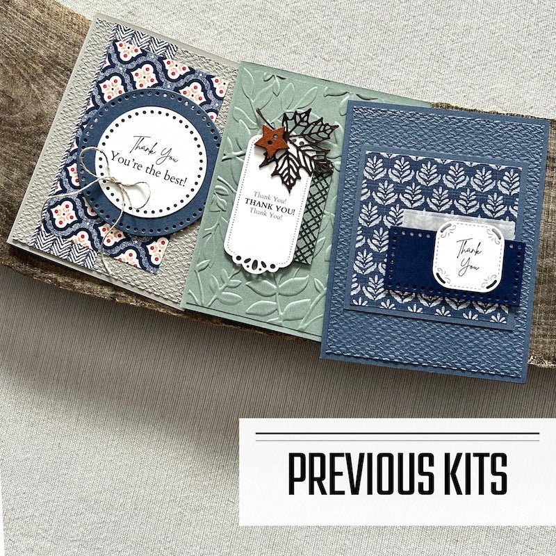 There are 3 finished cards, sitting on a wooden board, to show what cards have been sent in previous kits - The Craft Shoppe Canada