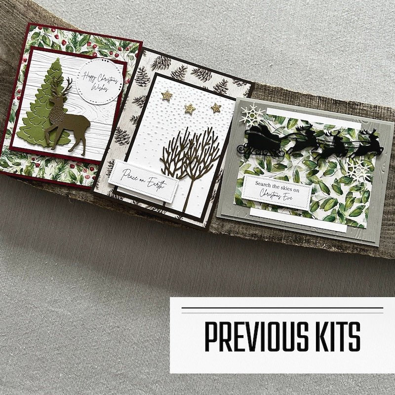 There are 3 finished cards, sitting on a wooden board, to show what cards have been sent in previous kits - The Craft Shoppe Canada