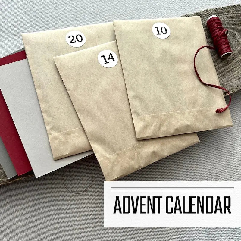 A DIY card making kit for 24 assorted greeting cards. This is an Advent Calendar featuring numbered packages from 1-24. There are 3 numbered kits sitting on a wooden board with a roll of red twine.