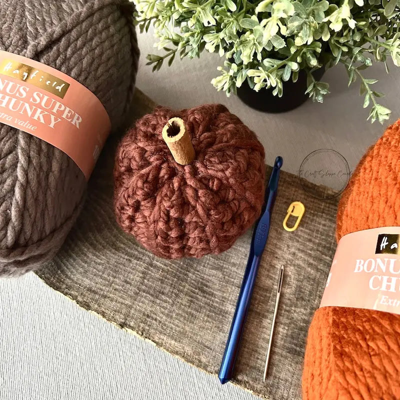 A finished pumpkin, made from this crochet kit, is sitting on a wooden board with yarn and a crochet hook.