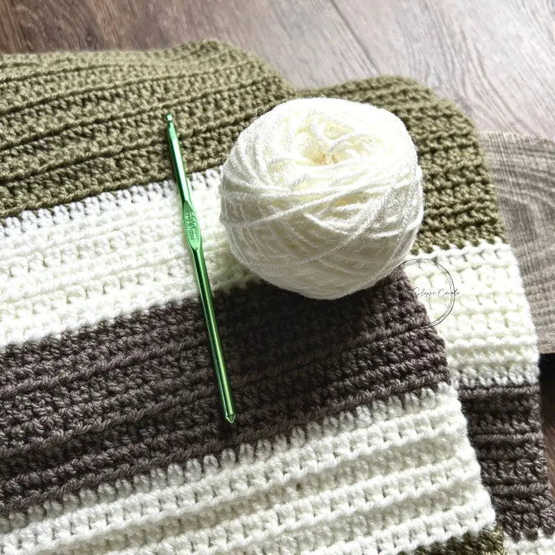 The finished baby blanket, made from this crochet kit, is lying on a wooden board with a ball of yarn and a crochet hook.