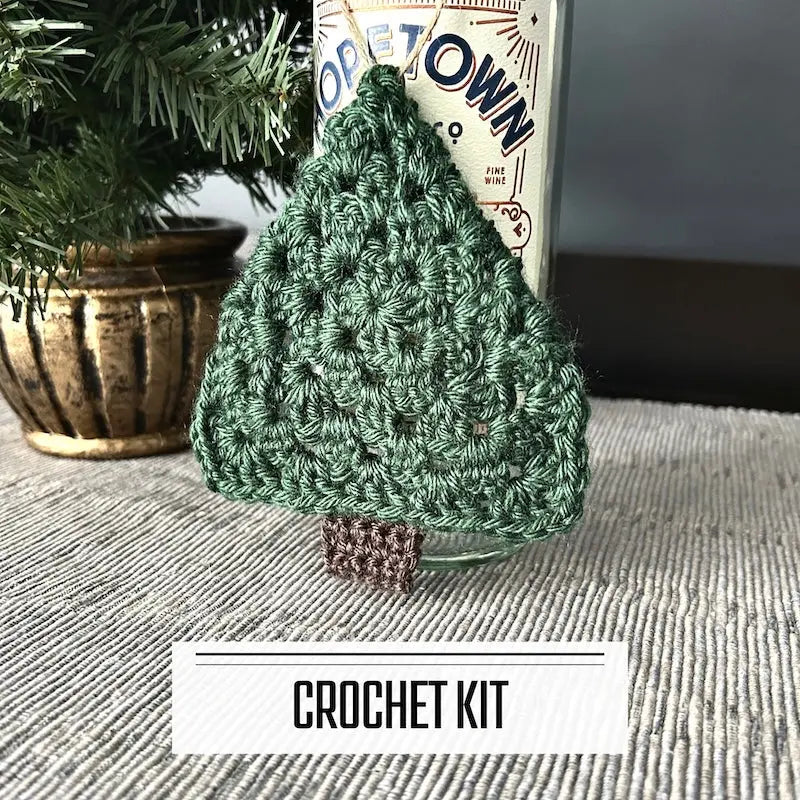 A DIY crochet kit for Christmas Tree Ornaments. The finished ornament is hanging over a bottle of wine, sitting on a table.