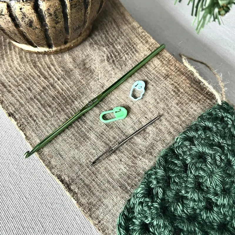 A crochet hook, 2 stitch markers and a tapestry needle are sitting on a wooden board.