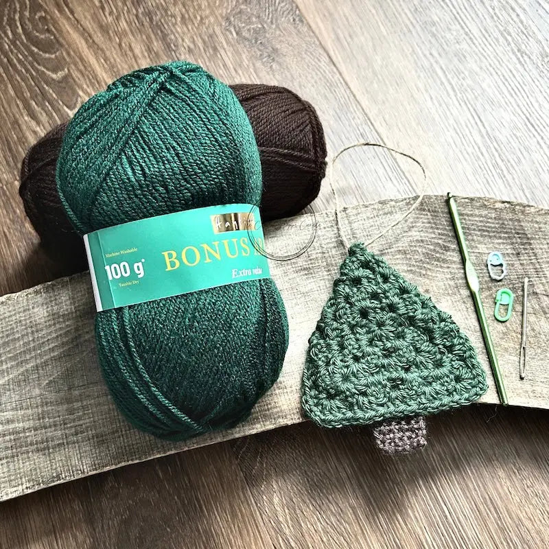 A ball of green and brown yarn, used in this crochet kit is sitting on a wooden board with a crochet hook, tapestry needle and 2 stitch markers.