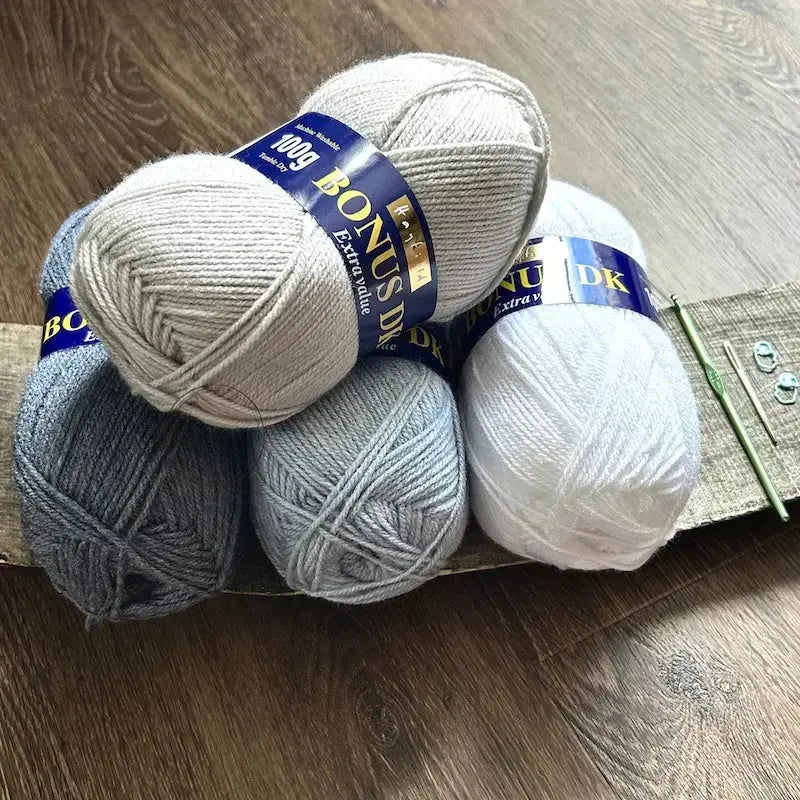 4 balls of yarn, that would be used in this crochet pattern, are sitting on a wooden board with a crochet hook, a tapestry needle and stitch markers.