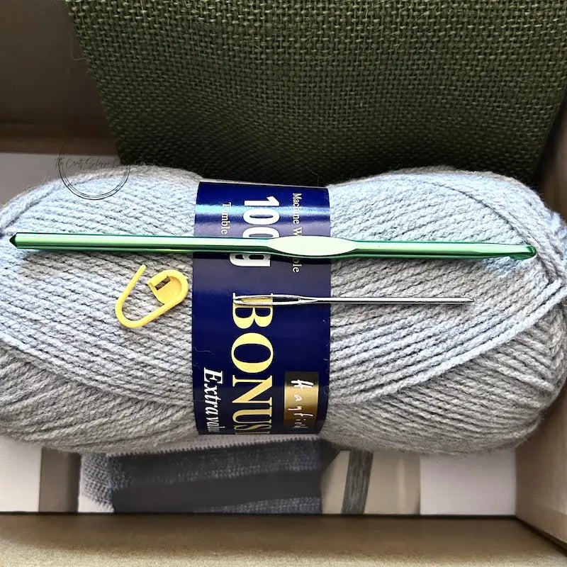 The yarn used in this crochet kit is lying in a box with a crochet hook, stitch marker and a tapestry needle.