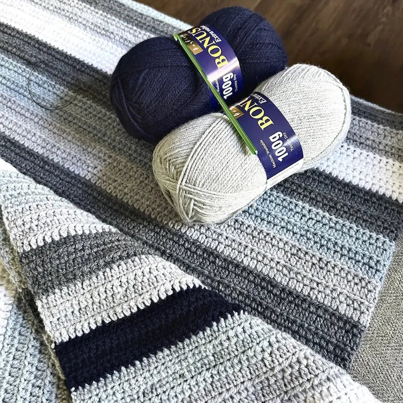 The finished couch throw, made from this crochet kit, is draped over a couch with 2 balls of yarn and a crochet hook.
