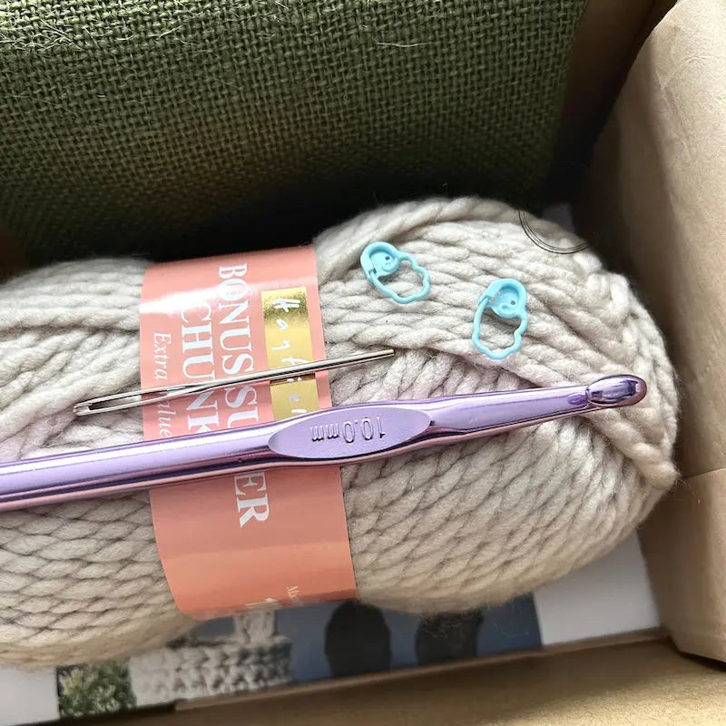 A ball of beige yarn is sitting in a box with a crochet hook, a tapestry needle and 2 stitch markers.