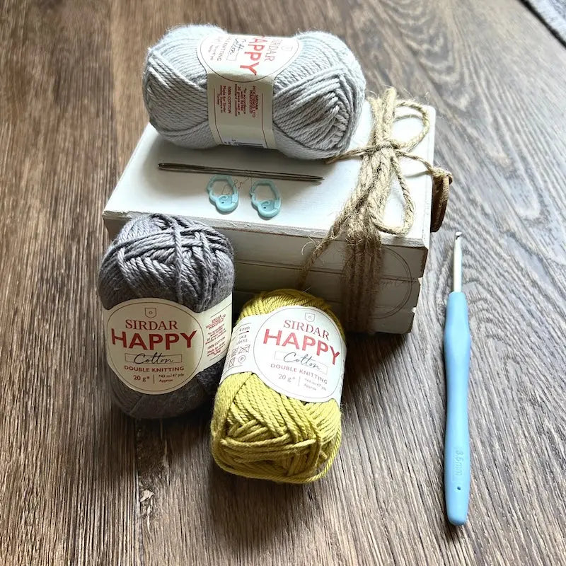 The balls of yard, used in this crochet kit, are sitting on a white wooden board with a crochet hook, tapestry and stitch markers.